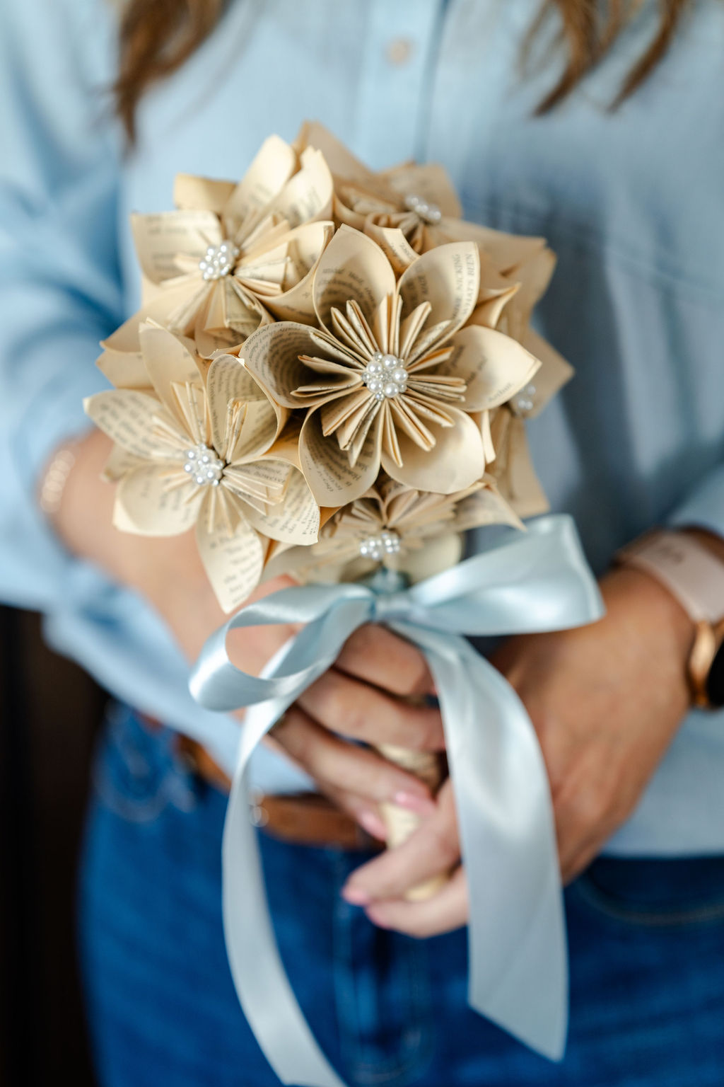 A woman is holding a bridal bouquet of origami paper flowers, diamantes and blue ribbon