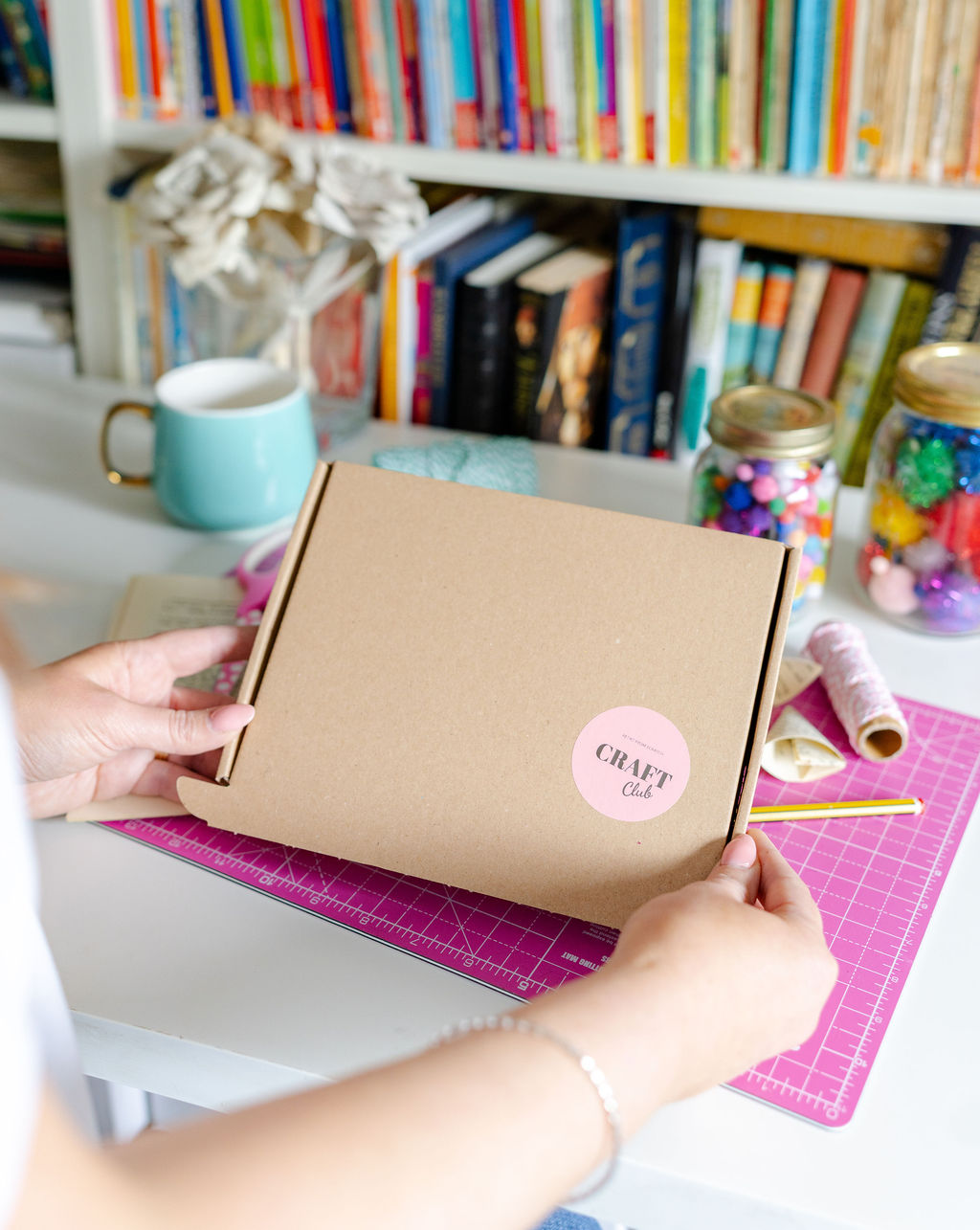 A woman is holding a brown box labelled 'Craft Club' with books on the shelf behind