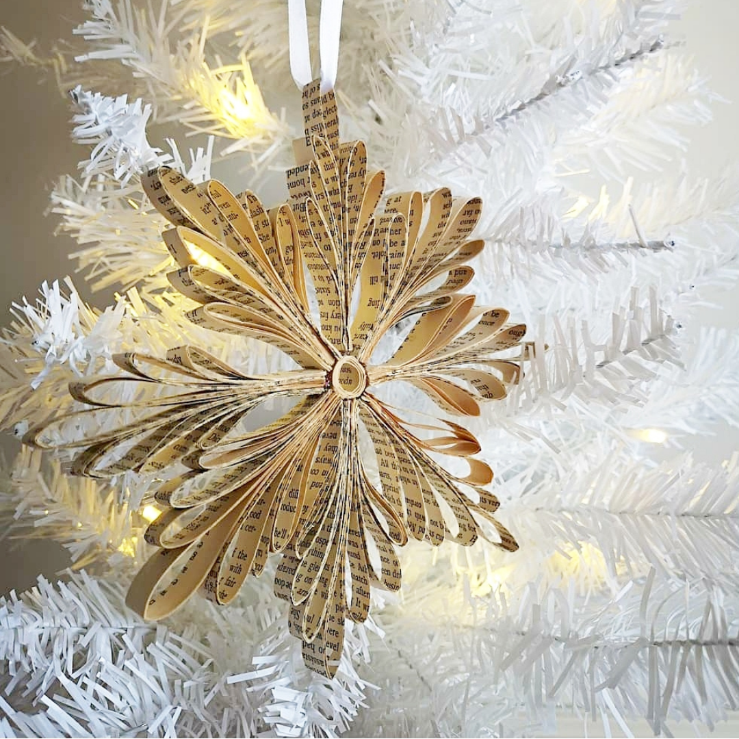 A snowflake made from strips of book paper hanging on a white Christmas tree